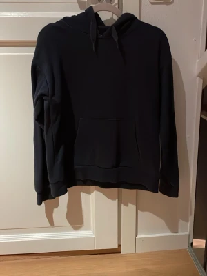 Svart hoodie med magficka - En stilren mörkblå hoodie med huva och dragsnören. Hoodien har en klassisk magficka framtill och ribbade muddar vid ärmslut och nederkant. Perfekt för en avslappnad och trendig look.