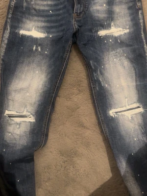 Blå slitna jeans Supply & Demand - Säljer ett par blå jeans från Supply & Demand med slitningar, distressed detaljer och vita färgstänk. Jeansen har en tvättad look och raka ben. Perfekta för dig som gillar en trendig och avslappnad stil. Inprincip helt nya