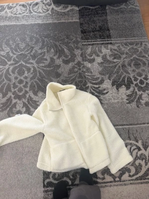 Vit teddyjacka från 157 - Mysig vit teddyjacka från 157 med hög krage och stora fickor framtill. Jackan har en fluffig och mjuk struktur i teddy-material som ger en avslappnad och trendig look. Perfekt för dig som vill ha en varm och stilren jacka till hösten eller vintern.