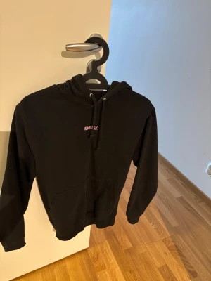 Svart hoodie SNJJK med rosa tryck - Svart hoodie från fröken snusk med huva och känguruficka. På bröstet står det snusk i rosa och på ryggen finns ett stort rosa tryck av en person på häst. Mjuk bomullskänsla och avslappnad passform, perfekt för en chill look. Använd 2 gånger