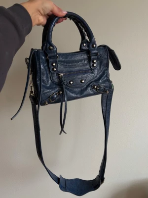 Mörkblå handväska - Säljer en mörkblå handväska i mjukt läder. Väskan har både handtag och en lång axelrem, silverfärgade nitar och dragkedja framtill. Klassisk city-modell. Grönt färgstänk som hamnat på axelremmen men går att ta bort axelremmen lätt. Sömmar som lossnat på handtaget men inget märkbart. 
