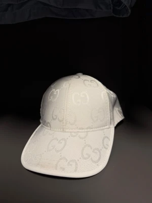 Vit keps från Gucci med GG-mönster - Säljer en stilren vit keps från Gucci med det klassiska GG-monogrammet över hela kepsen. Justerbar rem bak med gröna och röda detaljer. Tillverkad i canvasmaterial som ger en exklusiv känsla och passar perfekt till streetwear eller en clean look.