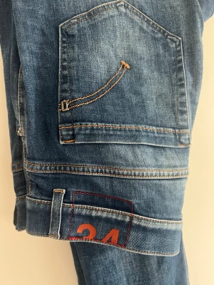 Blå dondup jeans  - Säljer ett par blå jeans med unik fickdetalj och orange kontrastsömmar. På benet finns en rödmarkerad ruta med siffran 24 i orange. Jeansen har en klassisk femficksdesign och är tillverkade i denim. Perfekta för dig som gillar streetwear och detaljer.