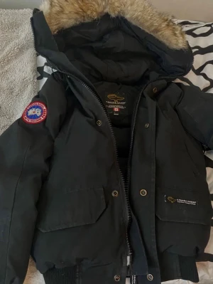 Svart pufferjacka från Canada Goose - Säljer en svart pufferjacka från Canada Goose med luva och fuskpälsdetalj. Jackan har ribbade muddar, två stora fickor framtill och klassisk Canada Goose-logga på ärmen. Perfekt för kalla vinterdagar och riktigt snygg streetstil. Bra för vintern och våren.