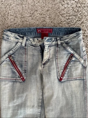 Ljusblå Miss Zhan jeans med röda dragkedjor - Unika ljusblå jeans från Miss Zhan jeans med snygga röda dragkedjor på fickorna och kontrastsömmar. Jeansen har en lätt utsvängd passform och cool tvättad look. Perfekta för dig som vill sticka ut med detaljer! Jeansen är dessutom stretchiga.         Innerbenslängden:83.  Midjemåttet:37.  