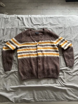 Brun kofta/cardigan med gulorange och vita ränder - Mysig brun kofta med knappar framtill och breda ränder i vitt, gulorange över bröstet och ärmarna. Koftan har v-ringning, lång ärm och ribbade muddar. Perfekt för lager på lager och en avslappnad stil.