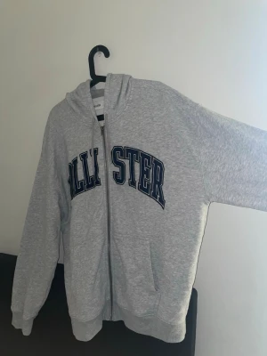 Grå zip hoodie från Hollister - Snygg grå hoodie med dragkedja från Hollister. Stor logga i mörkblått och vitt på bröstet. Mjuk bomullsblandning, huva med dragsko och två fickor framtill. Perfekt för chill dagar och enkel att matcha med jeans eller joggers. Storlek XL