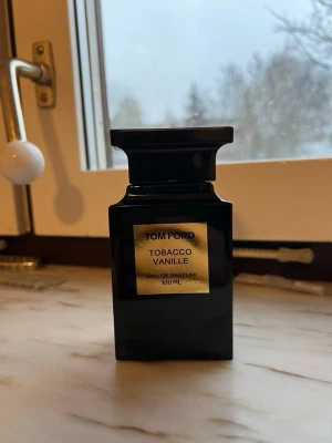 Tom Ford Tobacco Vanille 100 ml parfym - Tom Ford Tobacco Vanille Eau de Parfum i en elegant svart flaska med guldfärgad etikett och detaljer. Flaskan har en unik, fyrkantig form och rymmer 100 ml. Doften är känd för sina varma, kryddiga och söta toner av tobak och vanilj. Perfekt för dig som gillar exklusiva och lyxiga parfymer.