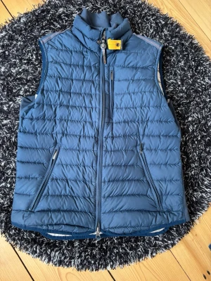 Blå dunväst från Parajumpers - Hej säljer en snygg blå dunväst från Parajumpers med ståkrage och dragkedja framtill. Västen har två fickor med dragkedja och en gul detalj vid halsen. Insidan är ljusgrå och materialet är lätt och quiltat för extra komfort. Är i väldigt bra skick, hör av er vid frågor eller funderingar 