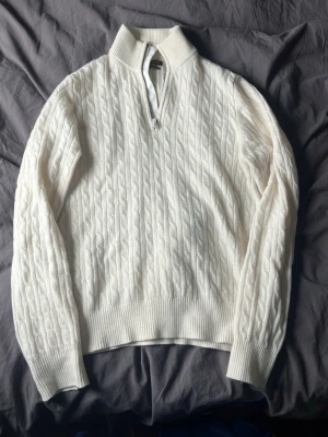 Vit kabelstickad tröja med halvzip - Kabelstickad vit tröja med halv dragkedja och hög krage. Tröjan har långa ärmar, ribbstickade muddar och nederkant. Perfekt för lager-på-lager och en clean look.
