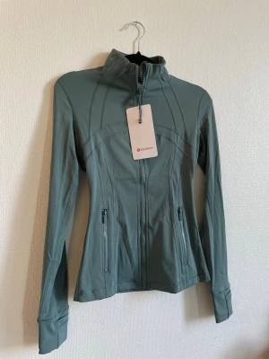 Grågrön träningsjacka från Lululemon - Slimmad träningsjacka från Lululemon i grågrön nyans. Jackan har hög krage, hel dragkedja framtill och två fickor med dragkedja. Tillverkad i stretchigt funktionsmaterial och har tumhål vid ärmsluten. Perfekt för träning eller powerwalks.