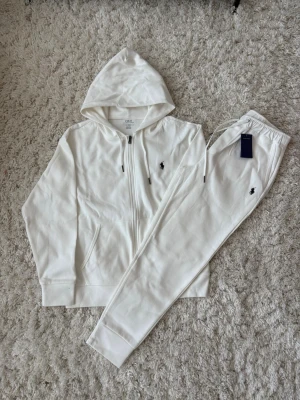 Vitt set från Polo Ralph Lauren - Snyggt vitt set från Polo Ralph Lauren med zip-hoodie och matchande byxor. Båda plaggen har den klassiska broderade loggan i svart. Hoodien har dragsko och fickor, byxorna har resår i midjan och dragsko. Mjukt och bekvämt material.