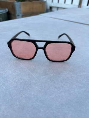 Svarta solglasögon med rosa glas - Snygga solglasögon med svart ram och stora, fyrkantiga rosa glas. Ramen har en cool detalj vid näsryggen och ger ett retroinspirerat intryck. Perfekt accessoar för dig som vill sticka ut med färgade glas och unik form. Väldigt snygga och perfekta till en varm sommardag 🤗🙌🍉