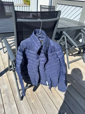 Mörkblå pufferjacka med hög krage - Snygg mörkblå pufferjacka med hög krage och horisontella sömmar. Jackan har dragkedja framtill och är lätt vadderad, perfekt för kalla dagar. Enkel och stilren design som passar till det mesta. Materialet är glansigt och ger en modern look.