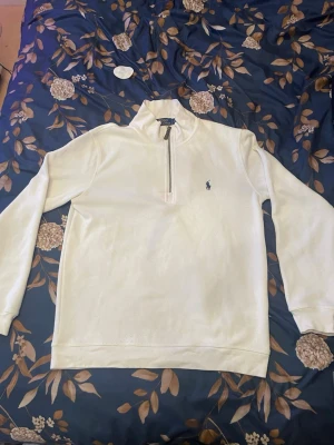 Vit half-zip sweatshirt från Polo Ralph Lauren - Vit sweatshirt med halv dragkedja från Polo Ralph Lauren. Klassisk design med broderad marinblå logga på bröstet och ribbade muddar. Perfekt för en clean och stilren look. Materialet är mjukt och bekvämt, passar till det mesta. Har små smink fläckar på som endast syns på nära håll. 