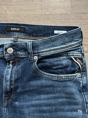 Blå jeans från Replay Wally's - Snygga blå jeans från Replay, modell Wally's, med klassisk femficksdesign och raka ben. Jeansen har slitningar och ljusare partier framtill för en trendig look. Tillverkade i denim med kontrastsömmar och metallknapp i midjan.