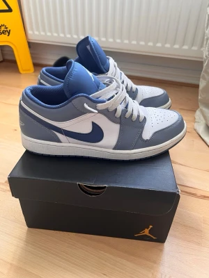 Air Jordan 1 Low blå/vit - Snygga Air Jordan 1 Low sneakers i blått och vitt läder med klassisk Nike Swoosh på sidan och Jumpman-logga på plösen. Skorna har vita snören, perforerad tåbox och en låg siluett som ger en clean och sportig vibe. Perfekta för dig som gillar streetwear och ikoniska modeller.