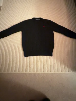 Mörkblå kabelstickad tröja Polo Ralph Lauren - Kabelstickad mörkblå tröja från Polo Ralph Lauren med rund halsringning och den klassiska röda logotypen broderad på bröstet. Tröjan har långa ärmar och ribbade muddar vid ärmslut och nederkant. Perfekt för en stilren och tidlös look.