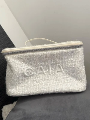 Vit necessär från CAIA - Snygg och rymlig vit necessär från CAIA med mjuk boucléstruktur och broderad logga framtill. Väskan har dragkedja och praktiskt handtag upptill. Perfekt för smink eller andra småsaker. Från tidigare julkalender 