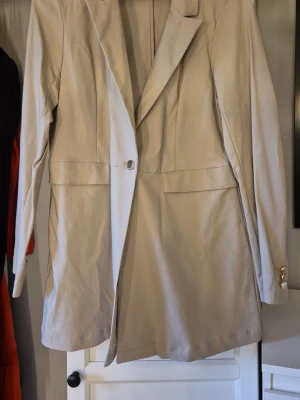 Beige lång kavaj med guldknappar - Stilren beige kavaj med lång passform och klassisk krage. Kavajen har två fickor framtill och stängs med en knapp. Ärmsluten är dekorerade med snygga guldknappar. Storlek 42 från Chiara Forthi. Mer Beige än vad bilden visar. Obs, katt finns i hemmet.