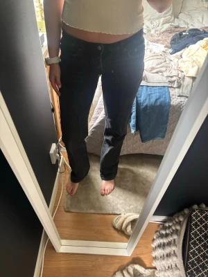 Svarta raka jeans med låg midja - Säljer ett par svarta jeans med rak passform och låg midja. Jeansen har klassisk femficksdesign och knappgylf. Perfekta för en avslappnad och trendig look. Materialet är denim med en mjuk känsla.