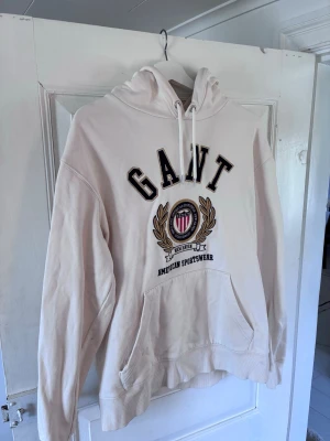 Vit hoodie från GANT med brodyr - Säljer en vit hoodie från GANT med stor broderad logga och emblem på bröstet. Klassisk känguruficka framtill, justerbar huva med snören och ribbade muddar. Tillverkad i mjuk bomullsblandning för skön känsla. Perfekt för en avslappnad och stilren look.