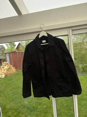 Svart overshirt från Asket - Snygg svart overshirt från Asket . med klassisk krage och tre stora fickor framtill. Rak passform och stängs med knappar. Perfekt att slänga över en t-shirt för en clean och stilren look. Gjord i ett slätt och tåligt material.