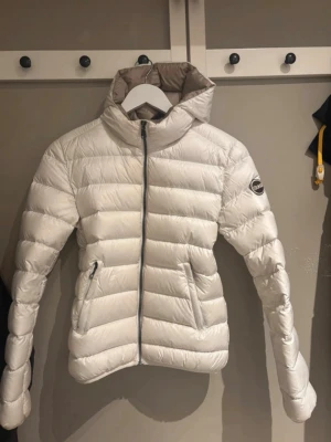 Colmar Jacka - Snygg vit dunjacka från Colmar med quiltad design och huva. Jackan har dragkedja framtill, två sidofickor med dragkedja och en beige insida. Märkeslogga på ärmen och modern passform. Perfekt för svala vår- och sommar dagar och för höst och vinter. Jackan tvättas en extra gång innan leverans och är i gott skick, endast använd under en vinter. 