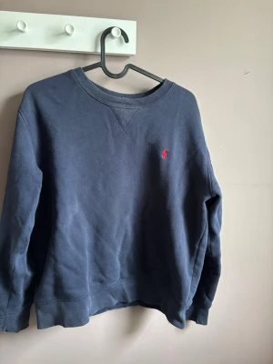 Mörkblå tröja från Polo Ralph Lauren - Mörkblå tröja från Ralph Lauren med klassisk rund halsringning och röd broderad logga på bröstet. Tröjan har långa ärmar och ribbade muddar vid ärmslut och nederkant. Perfekt för en stilren och avslappnad look. Storlek 14-16 men passar XS. Hur gärna av vid funderingar om pris eller annat!