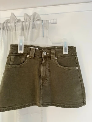 grön jeanskjol från stradivarius - Snygg olivgrön jeanskjol från stradivarius med klassisk femficksdesign, bälteshällor och knappstängning framtill. Kjolen är kort och har en rak passform, perfekt för en trendig och avslappnad look. Aldrig använd.