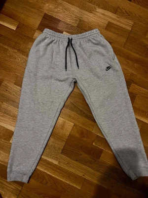 Gråa mjukisbyxor från Nike - Snygga grå mjukisbyxor från Nike med svart dragsko i midjan och klassisk Nike-logga på vänster ben. Byxorna har resår i midjan och vid bensluten för en skön passform. Perfekta för chill eller träning. Tillverkade i mjukt material.