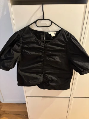 Svart puffärmad blus från H&M - Snygg svart blus från H&M med puffiga korta ärmar och rynkade detaljer framtill. Blusen har en rund halsringning och är tillverkad i ett lätt, glansigt material som ger en cool look. Perfekt att styla med jeans eller kjol.