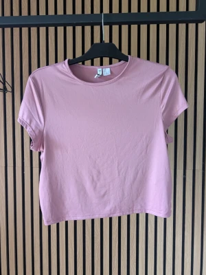 Rosa t-shirt från Divided - En stilren rosa kortare t-shirt från Divided med rund halsringning och korta ärmar. Perfekt för en avslappnad och trendig look. Tillverkad i mjukt material som känns skön mot huden. Ganska liten i storleken, stretchigt material Använd en gång på en babyshower