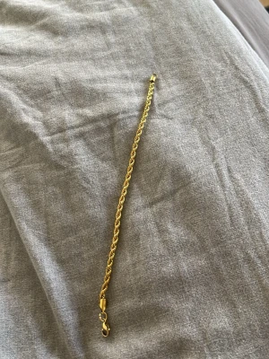 Guldfärgat kedjearmband - Snyggt armband i guldfärgad metall med tvinnad kedjedesign. Armbandet har ett klassiskt karbinlås och passar perfekt för att ge din outfit en extra touch av stil. Enkel men ändå iögonfallande design som funkar till många olika looks.