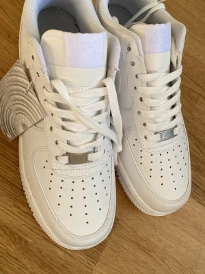 Nike Air Force 1 vita sneakers - Klassiska Nike Air Force 1 sneakers i helvitt läder med perforerad tå, vita skosnören och diskret Nike-logga på plösen. Snygg och tidlös modell med låg profil och metallbricka på snörningen. Perfekta för en clean och stilren look. Skriv vid minsta fråga 💖💖