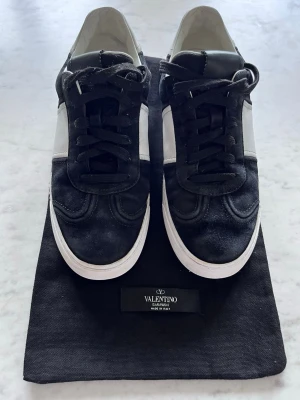 Svarta Valentino flycrews - Snygga svarta sneakers från Valentino med vita detaljer och vit sula. Skorna har klassisk låg modell, svarta skosnören och coola nitar på baksidan av sulan. Tillverkade i mocka och läder för en lyxig känsla. Perfekta för dig som gillar exklusiva streetwear-vibbar.