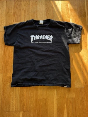 Svart Thrasher t-shirt M - Klassisk svart Thrasher t-shirt med vit logga och texten 'SKATEBOARD MAGAZINE' på bröstet. Rund halsringning och korta ärmar. Perfekt för dig som gillar streetwear och skatekultur.