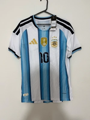 Argentina matchtröja Adidas nr 10 - Säljer en officiell Argentina matchtröja från Adidas med klassiska blå och vita ränder, guldiga detaljer och nummer 10 på bröstet. Tröjan har FIFA World Champions 2022-märke och AFA-logga. Tillverkad i lätt och ventilerande material, perfekt för fotboll eller träning.