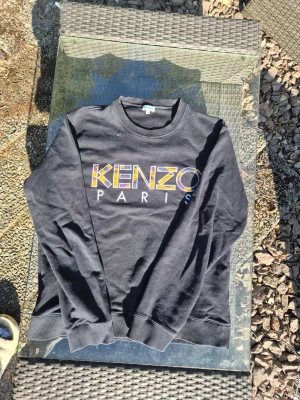 Svart Kenzo Paris sweatshirt - Snygg svart sweatshirt från Kenzo med färgglatt broderat Kenzo Paris-tryck på bröstet. Tröjan har rund halsringning, långa ärmar och ribbade muddar vid ärmslut och nederkant. Perfekt för en trendig och avslappnad stil. Sitter som en m