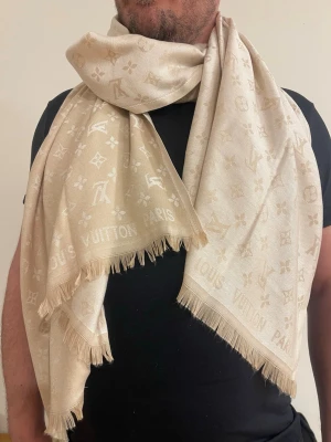 Beige  Vuitton halsduk - Stilren beige halsduk från  Vuitton med ikoniskt monogrammönster och fransade kanter. Mjuk och lyxig känsla, perfekt att svepa runt halsen för en trendig look. Diskret logotyp och klassisk design som lyfter vilken outfit som helst.