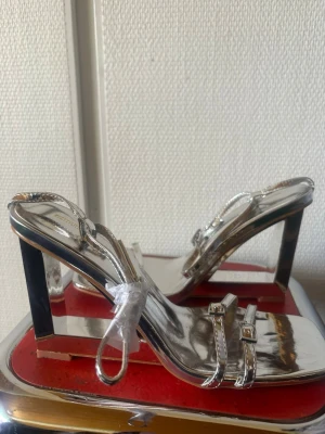 Silvriga pumps med fyrkantig tå - Snygga silvriga pumps med fyrkantig tå och bred klack. Skorna har tunna remmar över foten och runt ankeln, vilket ger en trendig och modern look. Perfekta för dig som vill sticka ut med metallic och spegelblank finish. Passar till både jeans och klänning.