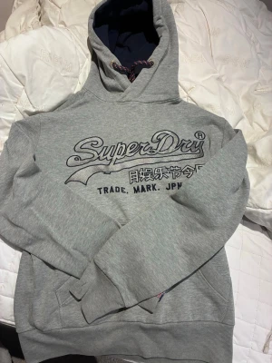 Grå Superdry hoodie med tryck - Snygg grå hoodie från Superdry med stort broderat logotyptryck på bröstet och japanska tecken. Mörkblå insida i huvan och dragsko med mönstrade snören. Perfekt för en avslappnad streetstyle-look.