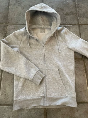 Ljusgrå zip-hoodie från Cubus - En stilren ljusgrå hoodie med dragkedja från märket Cubus. Hoodien har huva med dragsko, två fickor framtill och ribbade muddar vid ärmslut och nederkant. Perfekt för en avslappnad look och enkel att matcha med andra plagg.