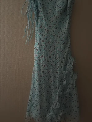 Ljusblå prickig klänning Milady - Supersöt ljusblå långklänning från Milady med prickigt mönster och volangdetaljer längs sidan. Klänningen har smala axelband och är tillverkad i ett lätt, flowy material som känns perfekt för varmare dagar.