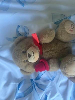 Söt nallebjörn med röd ros och text nedan - Mysig brun nallebjörn i mjuk plysch med en röd rosett runt halsen och en röd tygros i handen. På ena tassen står det 'I ♥ U' broderat med röd tråd. Perfekt som present till någon du tycker om.