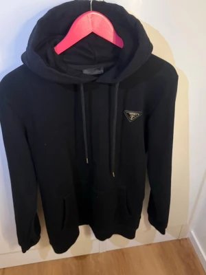 Svart hoodie från Prada med logga - Snygg svart hoodie/Prada  med klassisk huva och dragsko. På bröstet sitter en ikonisk trekantig Prada-logga i metall. Hoodien har känguruficka framtill och ribbade muddar. Perfekt för en stilren och trendig look.