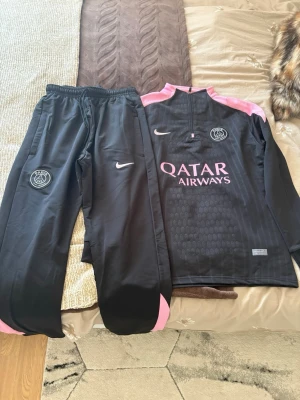 Svart PSG träningströja från Nike - Svart långärmad träningströja från Nike och Paris Saint-Germain med rosa detaljer och halv dragkedja. PSG-logga på bröstet och Qatar Airways-tryck framtill. Perfekt för dig som gillar sportig stil och fotbollsinspirerat mode.