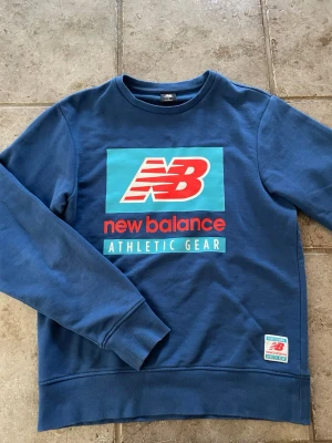Blå sweatshirt från New Balance - Snygg blå sweatshirt från New Balance med stort tryck på bröstet i rött, vitt och turkost. Tröjan har rund halsringning, långa ärmar och ribbade muddar. Perfekt för en sportig och avslappnad stil. Materialet är mjukt och bekvämt, troligen bomullsmix.