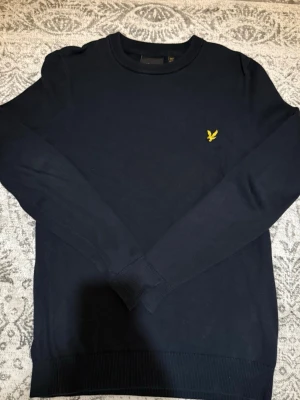 Mörkblå tröja från Lyle & Scott - Snygg mörkblå tröja från Lyle & Scott med rund halsringning och klassisk gul logga på bröstet. Tröjan har långa ärmar och ribbade muddar vid ärmslut och nederkant. Perfekt för en stilren och enkel look.