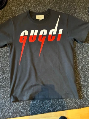 Svart Gucci t-shirt med röd/vit logga - snygg svart t-shirt från Gucci med stor, iögonfallande logga i vitt och rött framtill. T-shirten har klassisk passform och rund hals. Perfekt för dig som vill sticka ut med en trendig och exklusiv look.